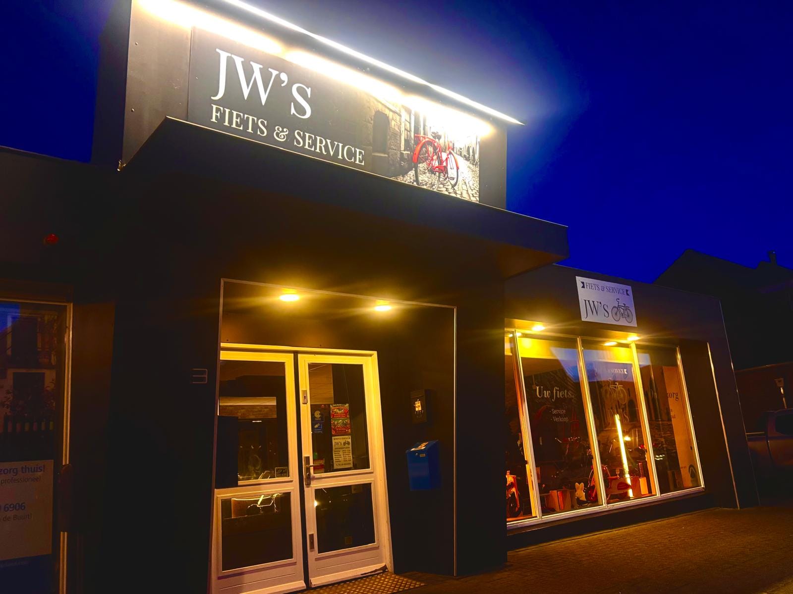 Jw's Fiets & Service winkel bij avond, verlicht uithangbord